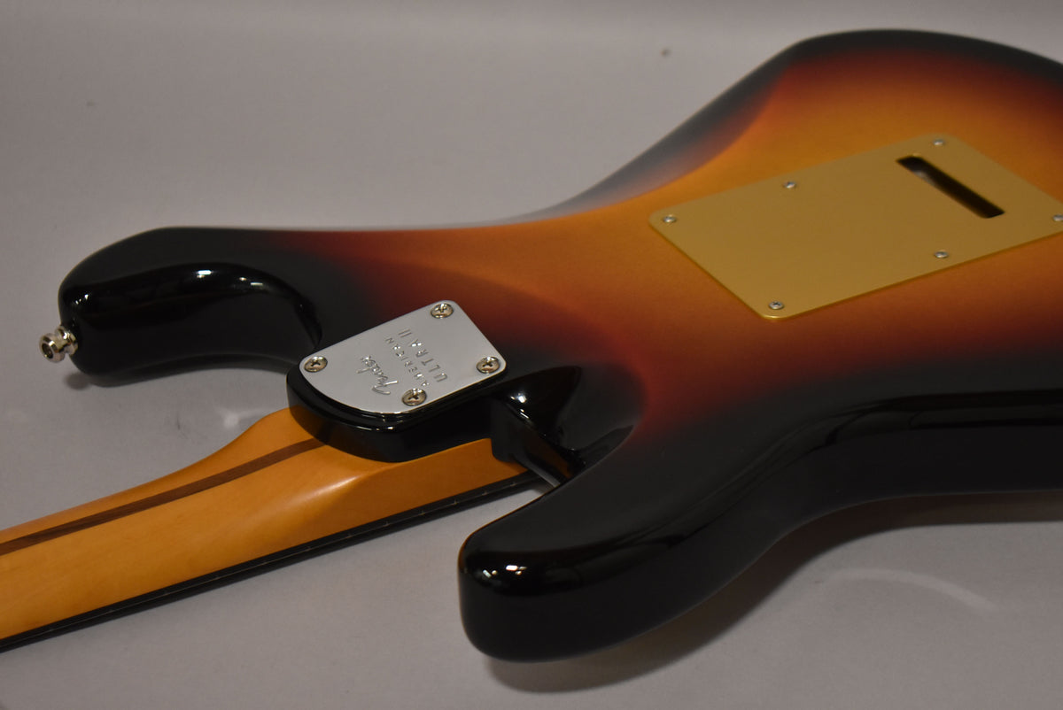 The guitar's back shows a glossy ultraburst finish, a gold tremolo cover, and a chrome neck plate with a rounded heel.