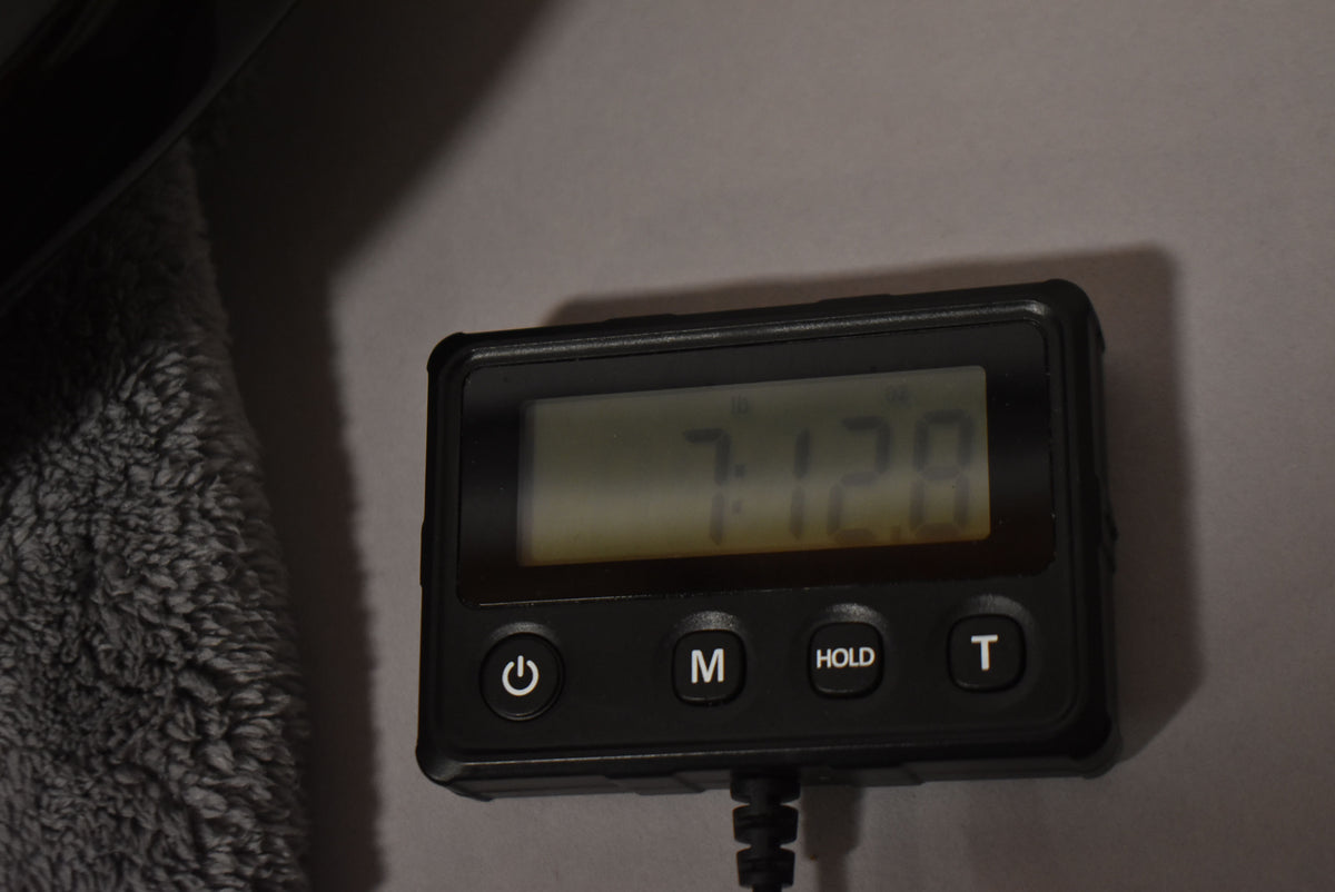 A black digital meter with a display showing "7.128" and buttons for power, M, HOLD, and T, next to a gray fuzzy material.