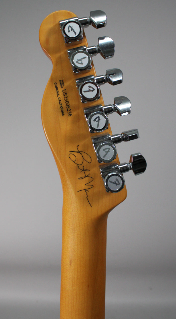 The guitar's light brown headstock and neck are angled, showing six chrome tuners and a black signature on the headstock.