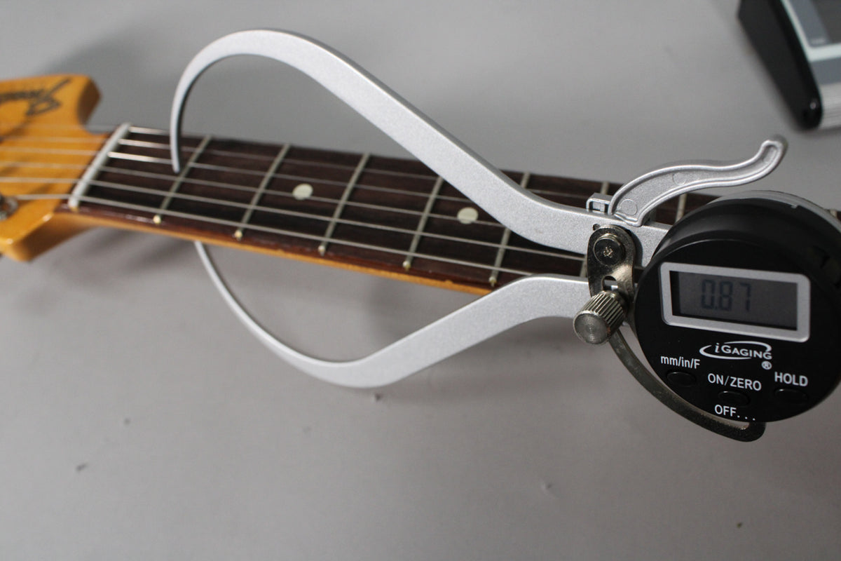 A digital caliper measures the guitar's fret height at 0.87mm, showing the dark fretboard and light-colored headstock.