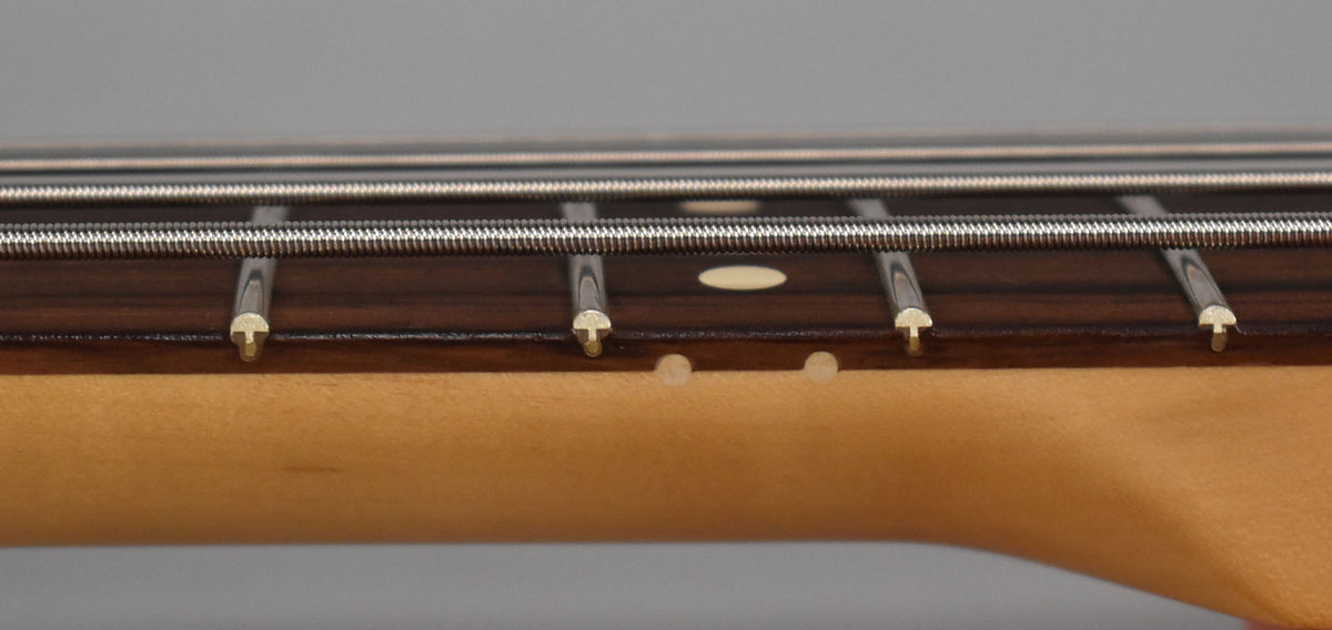 Close-up of the bass neck from the side, showing the dark fretboard, light maple neck, frets, strings, and two small dings.