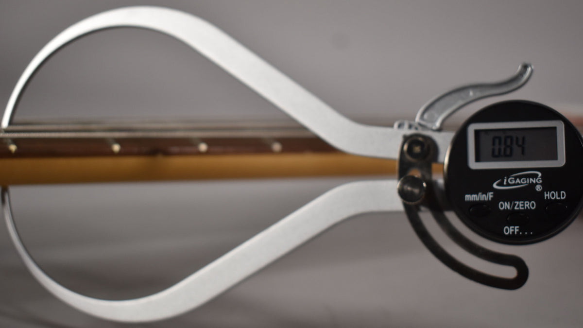 A digital caliper measures the bass's neck thickness at 0.84 inches, showing the fretboard and strings from above.