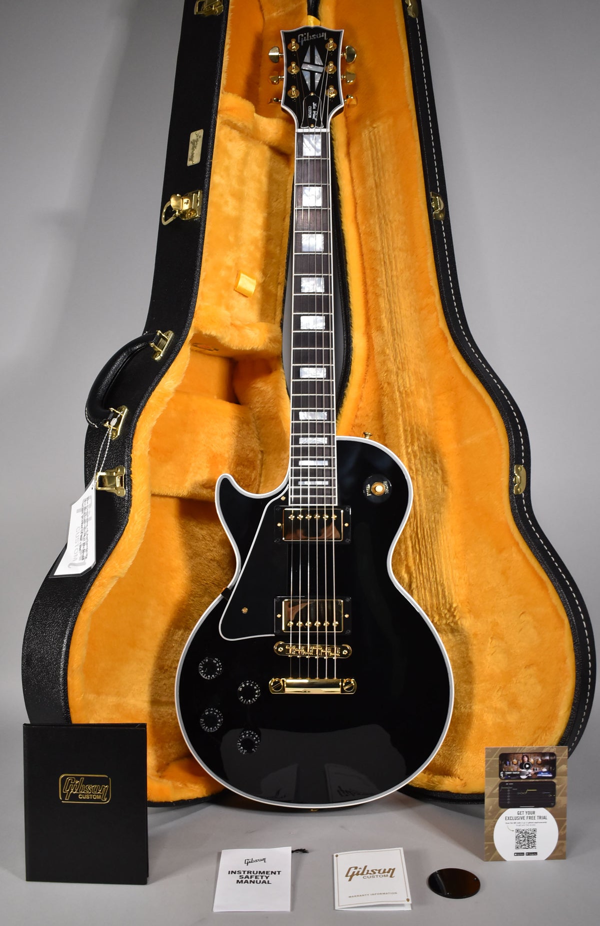 Full view of a glossy black Les Paul Custom with gold hardware, white binding, and block inlays, resting in its open, orange-lined hard case.