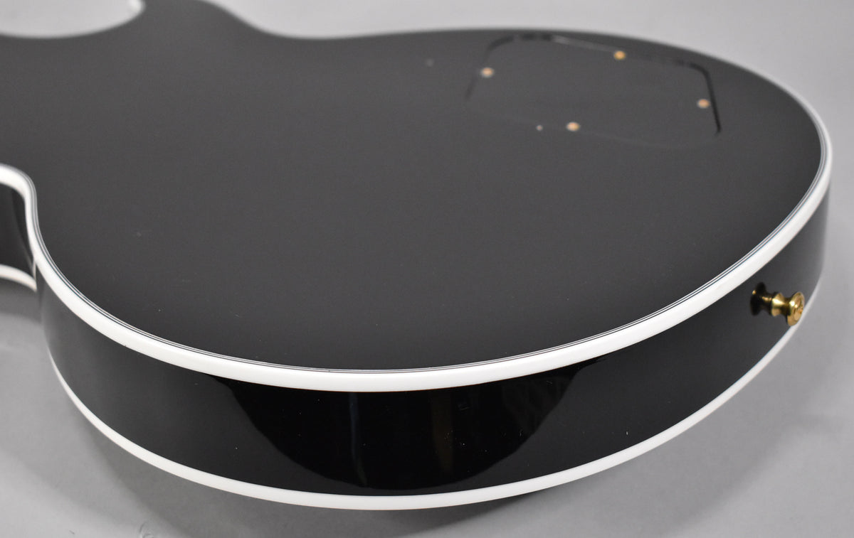 The guitar's lower bout, showing the glossy ebony back and side with white binding, and a gold strap button.