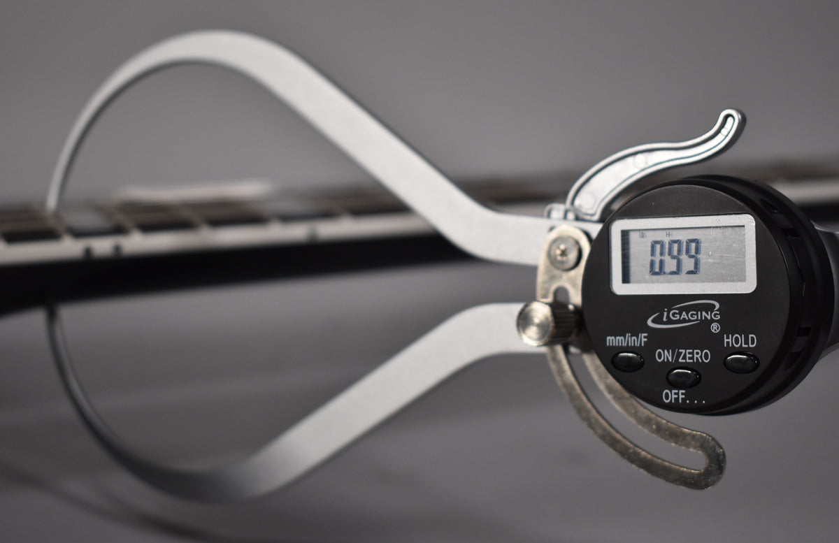 A digital caliper measures the guitar's fretboard, showing a reading of 0.99, with the gloss ebony finish visible.