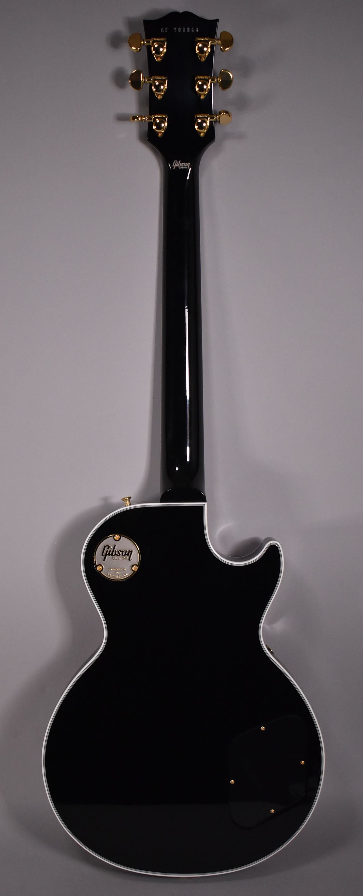 The back of a glossy black electric guitar with white binding, gold tuners, and a gold medallion on the upper bout.