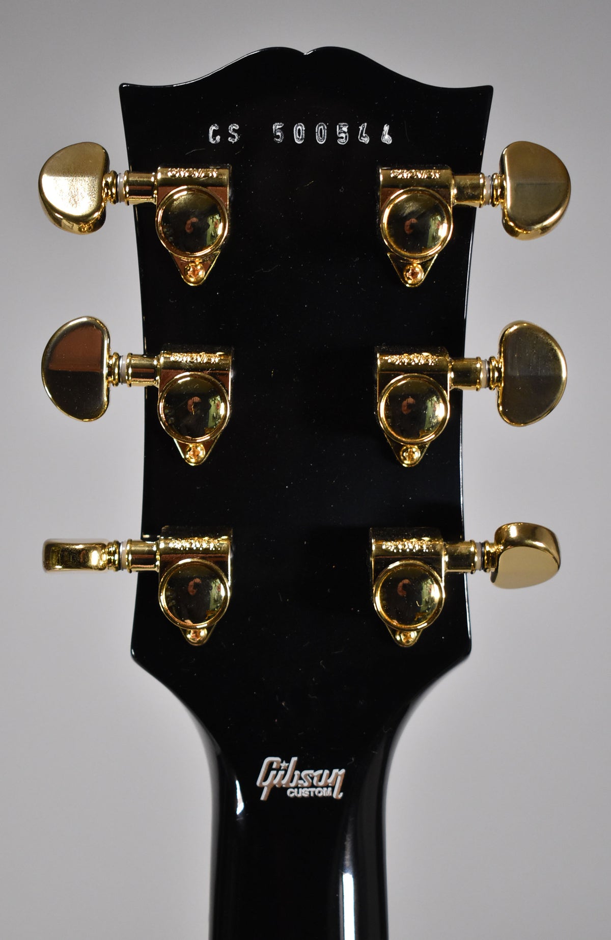 The back of a glossy black headstock with six gold tuning machines, three on each side, and a white Gibson Custom logo.