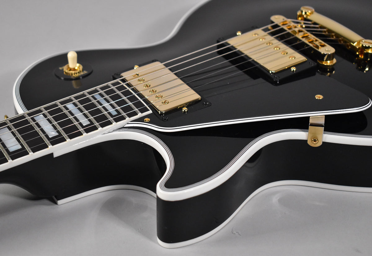 Close-up of a glossy black Les Paul Custom, showing the upper body, gold hardware, pickguard, and part of the fretboard.