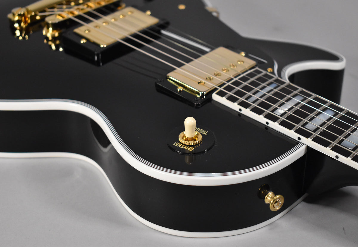 Close-up of a glossy black guitar body with white binding, showing gold hardware, a cream pickup selector switch, and part of the fretboard.
