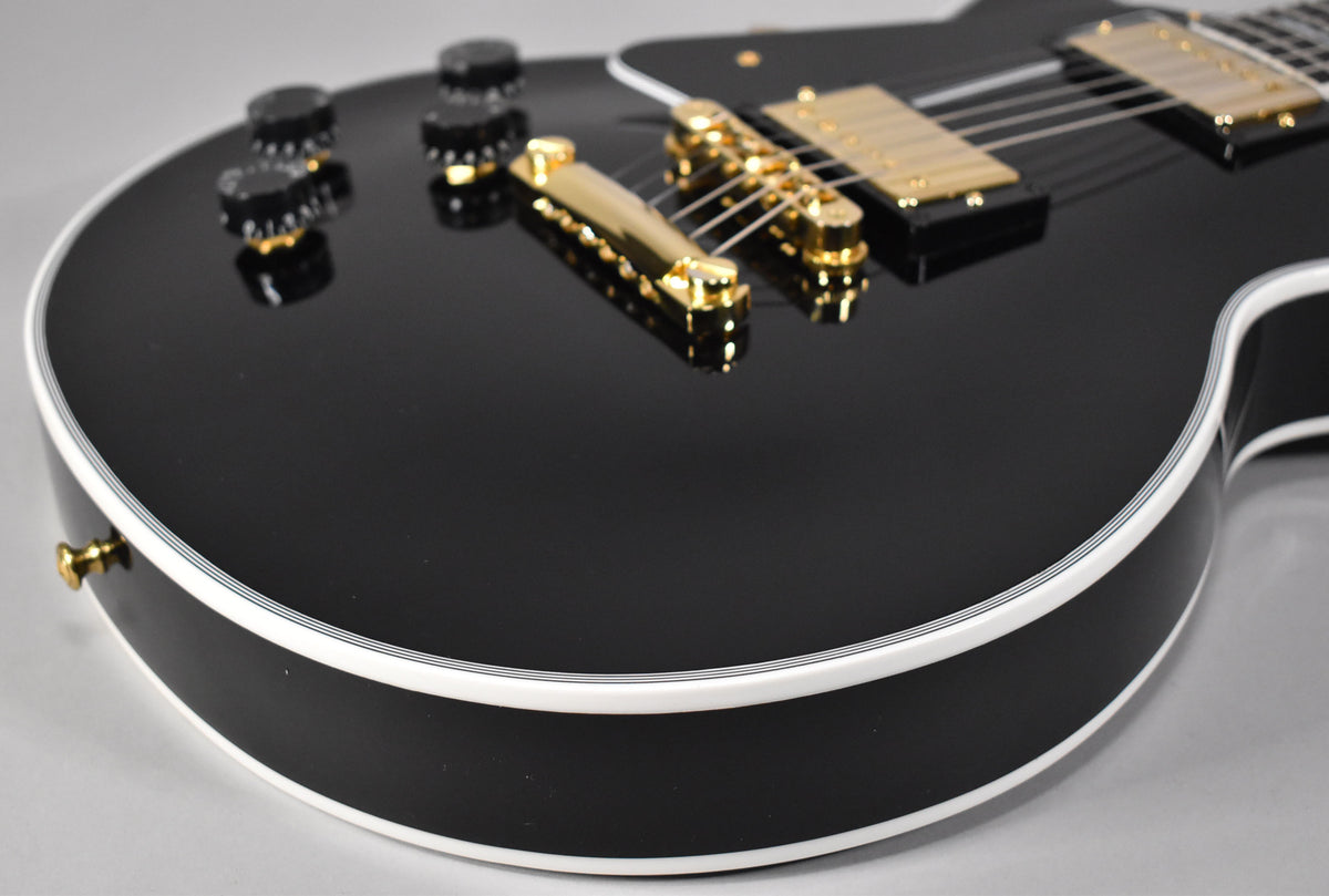 Close-up of the gloss black body with white binding, gold bridge, tailpiece, and humbucker pickups, and four black control knobs.