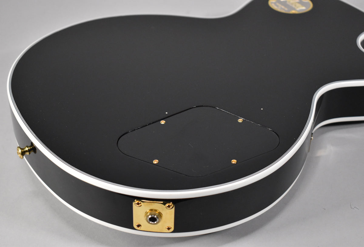 The guitar's gloss black body, with white binding, is angled to show the gold output jack and control cavity cover.