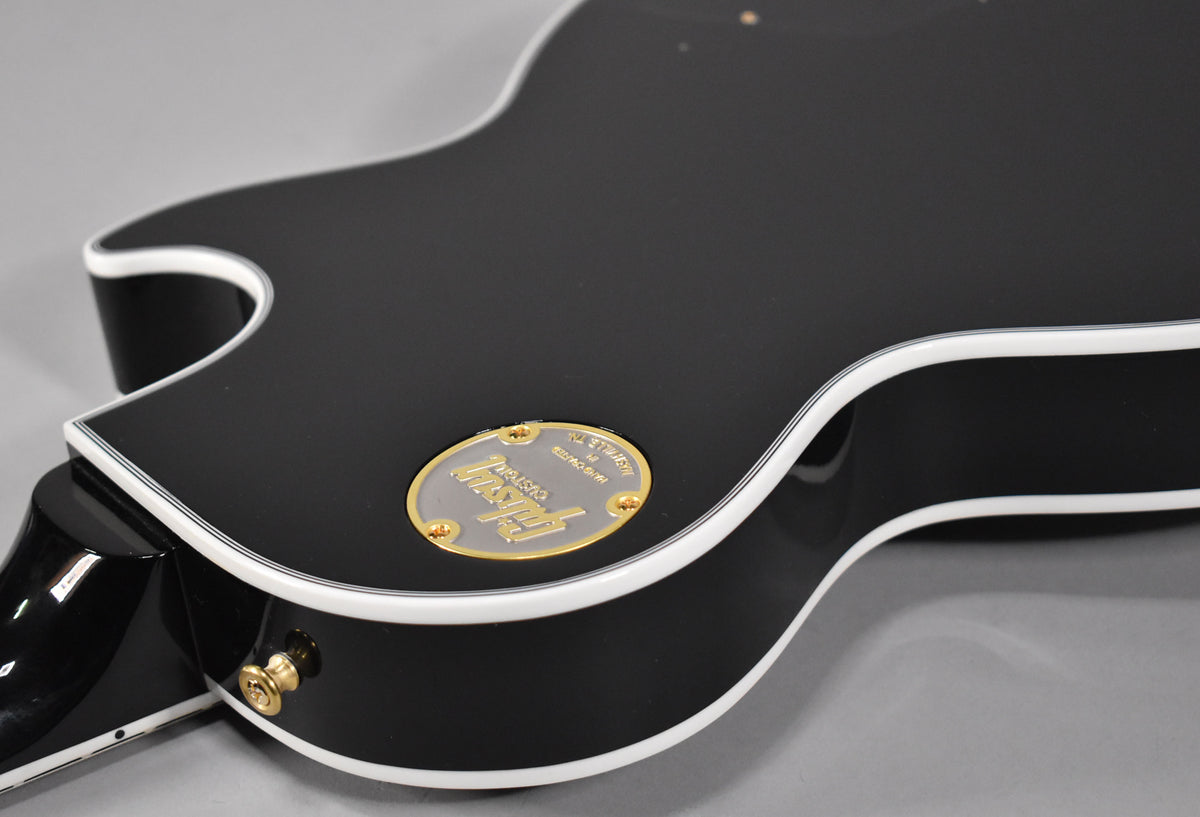 The guitar's gloss black body, with white binding, is shown from the back, angled to reveal the gold output jack and a gold-on-silver backplate.