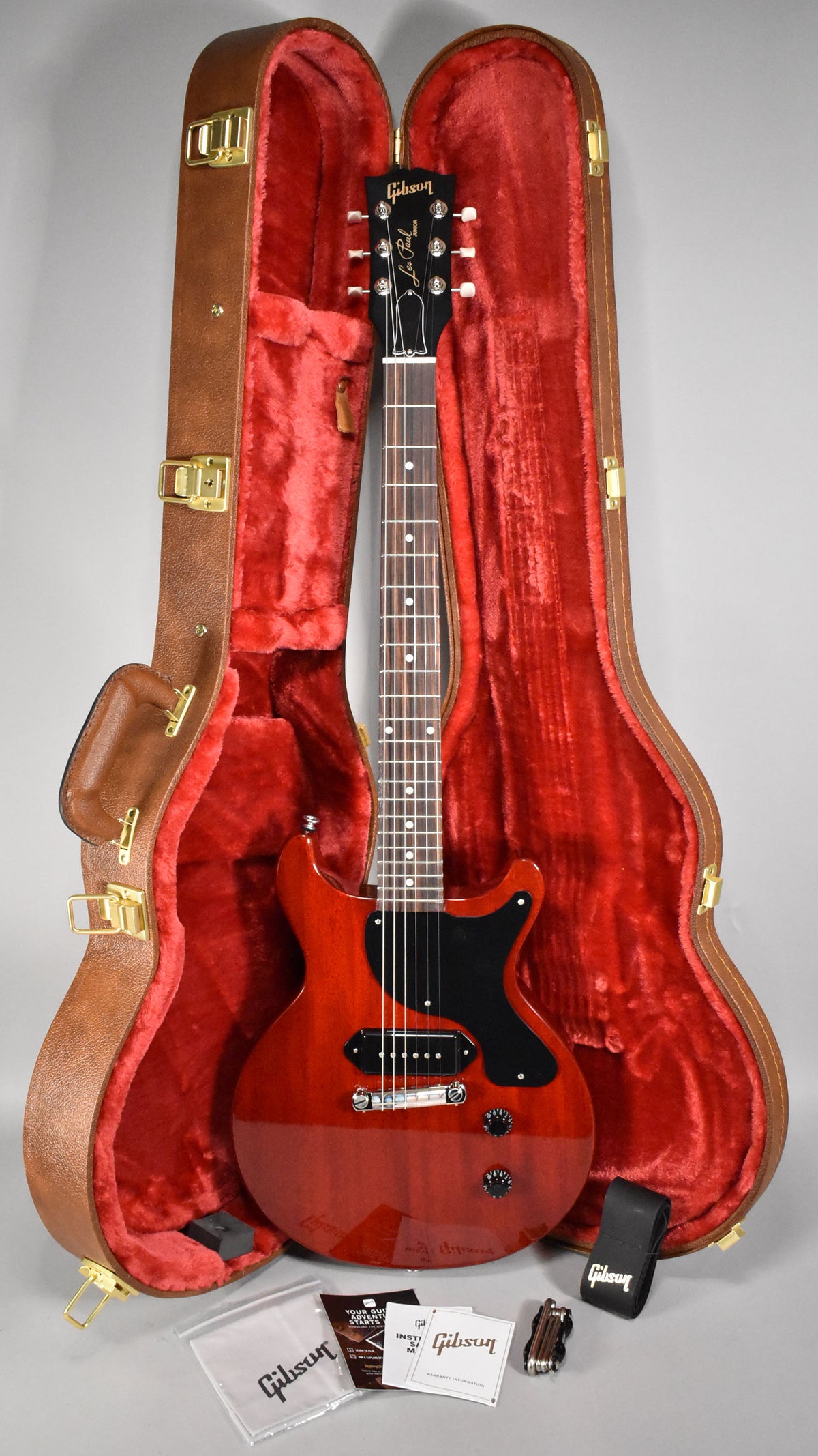 Full-length view of a glossy cherry red double-cut electric guitar with a black pickguard, nestled in a brown hard case with red plush lining.