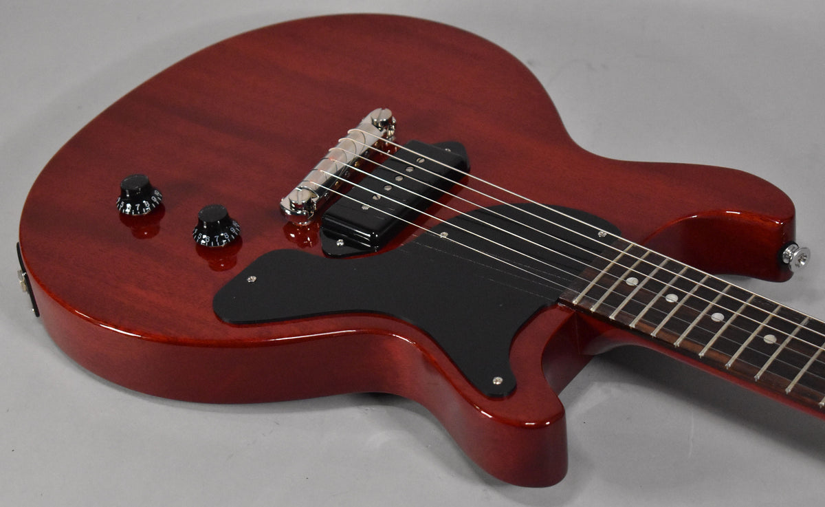 The cherry red double-cutaway body of the guitar is angled from the upper left, showing the black pickguard, P90 pickup, and two control knobs.