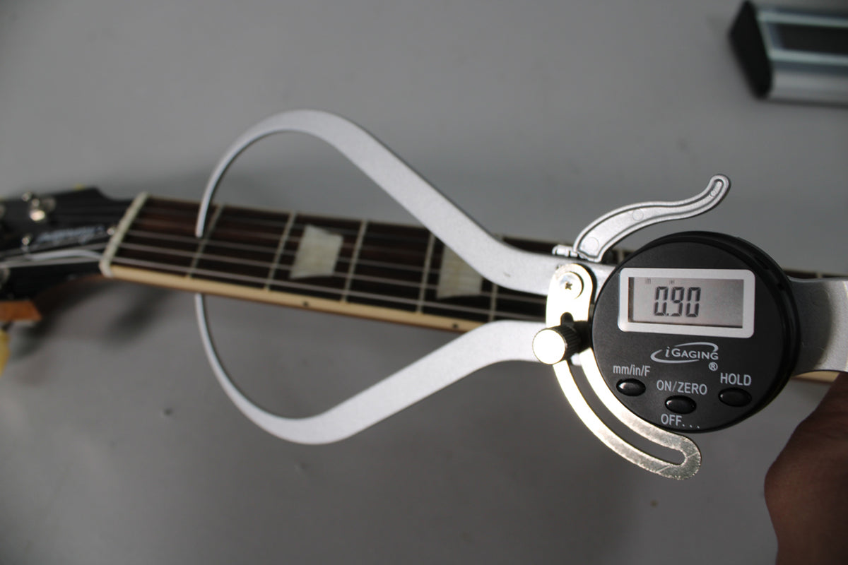 A digital caliper measures the fretboard, showing 0.90 on its screen, with trapezoid inlays visible on the dark wood.