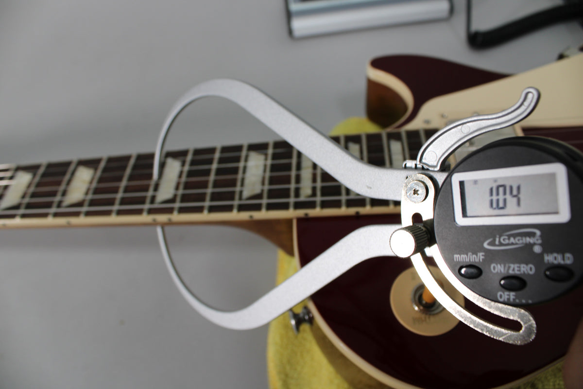 A digital caliper measures the guitar's fretboard, showing trapezoid inlays and a dark burgundy body with cream binding.