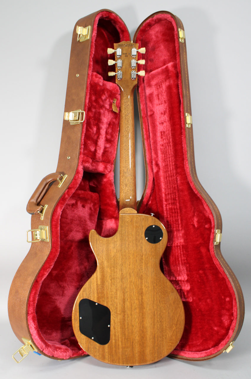 The back of a Les Paul guitar with a natural wood finish and black control cavity covers, resting in a brown case with red plush lining.