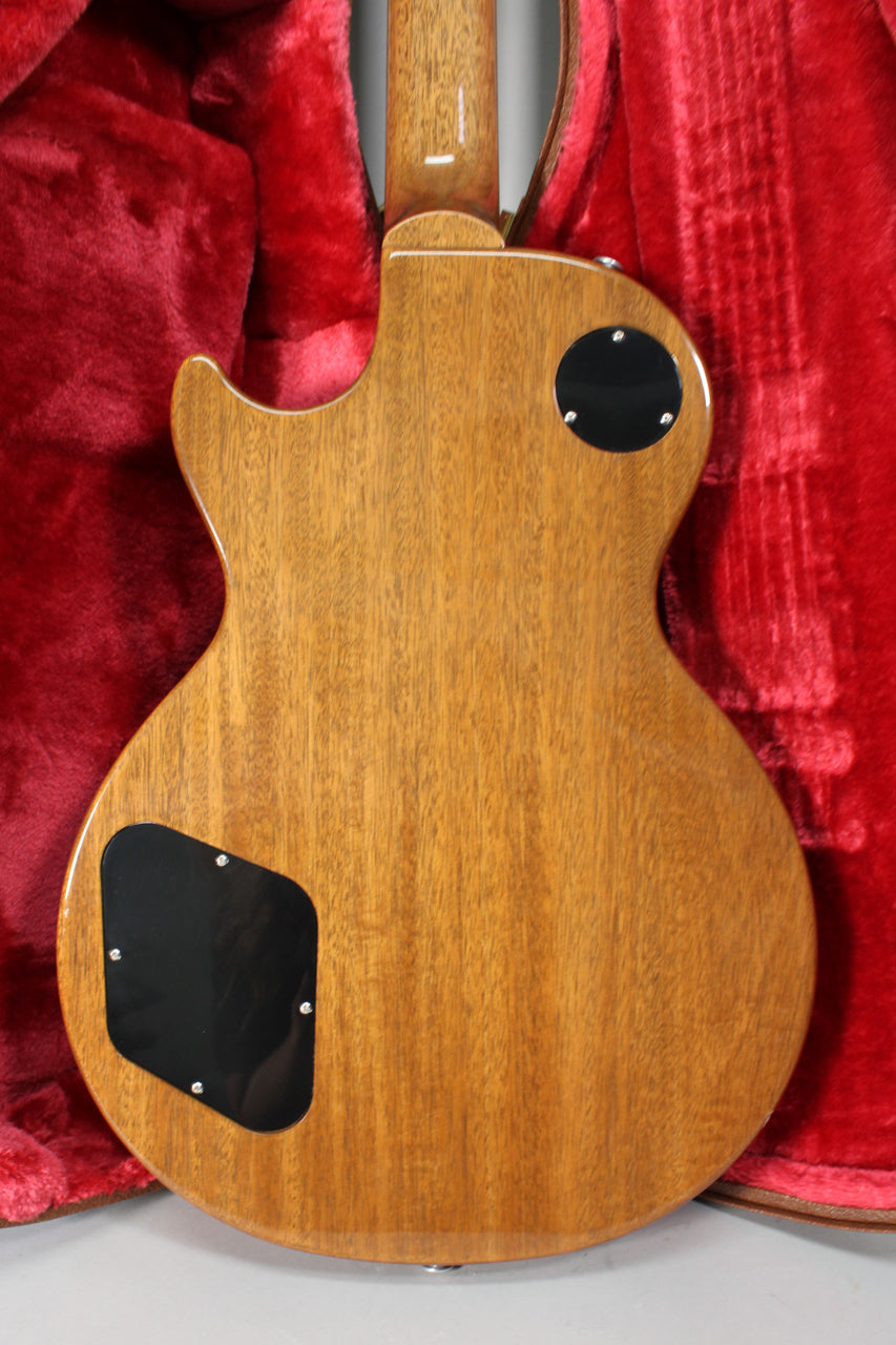 The back of a Les Paul guitar with a natural wood finish, showing two black control cavity covers, resting in a red plush case.