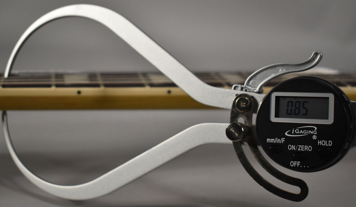 A digital caliper measures the guitar's neck, showing a reading of 0.85, with frets and dot inlays visible.