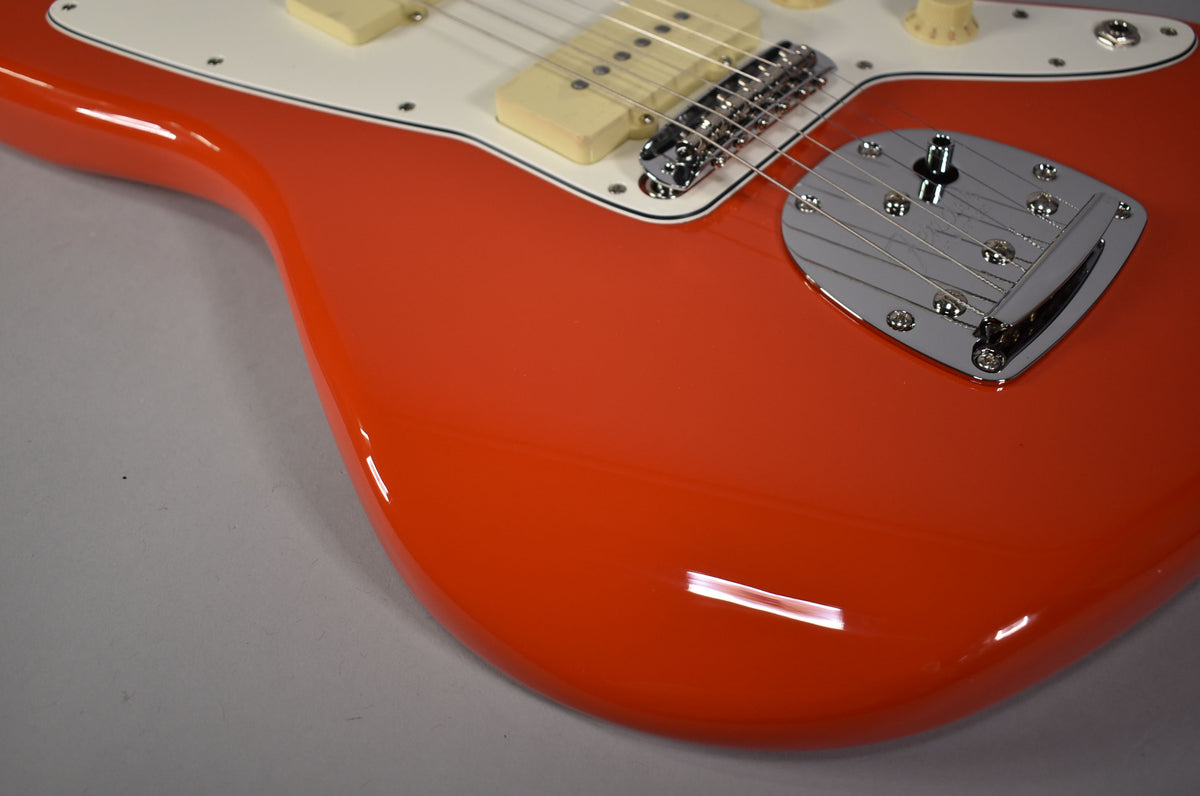 The lower body of a glossy coral red Jazzmaster, showing the white pickguard, cream pickups, chrome bridge, and vibrato unit.