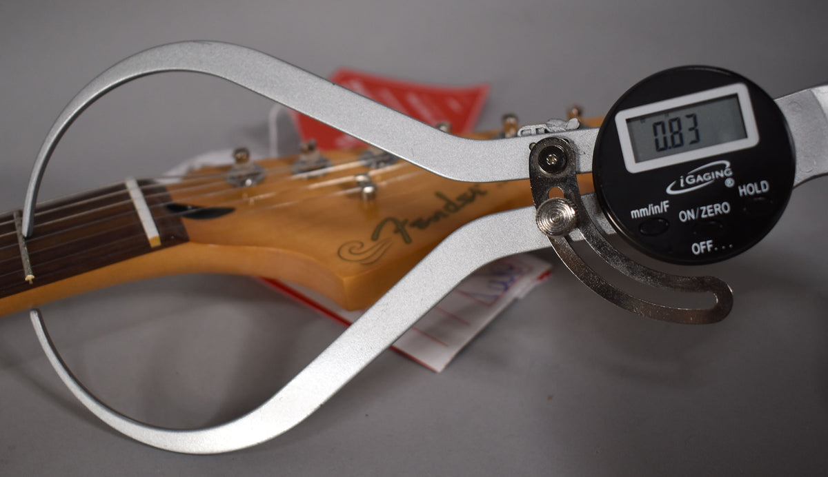 Digital calipers measure the guitar's headstock and nut, displaying "0.83" on the screen, with tuning pegs visible.