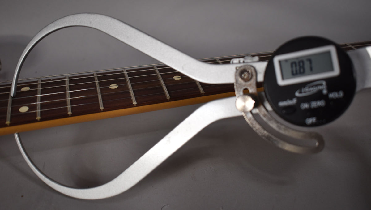 A digital caliper measures the guitar's fretboard, displaying "0.87" on its screen.