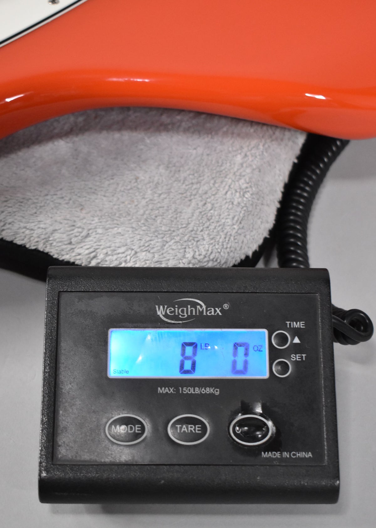The guitar's coral red body rests on a gray towel, with a digital scale displaying "8 LB 0 OZ" in the foreground.