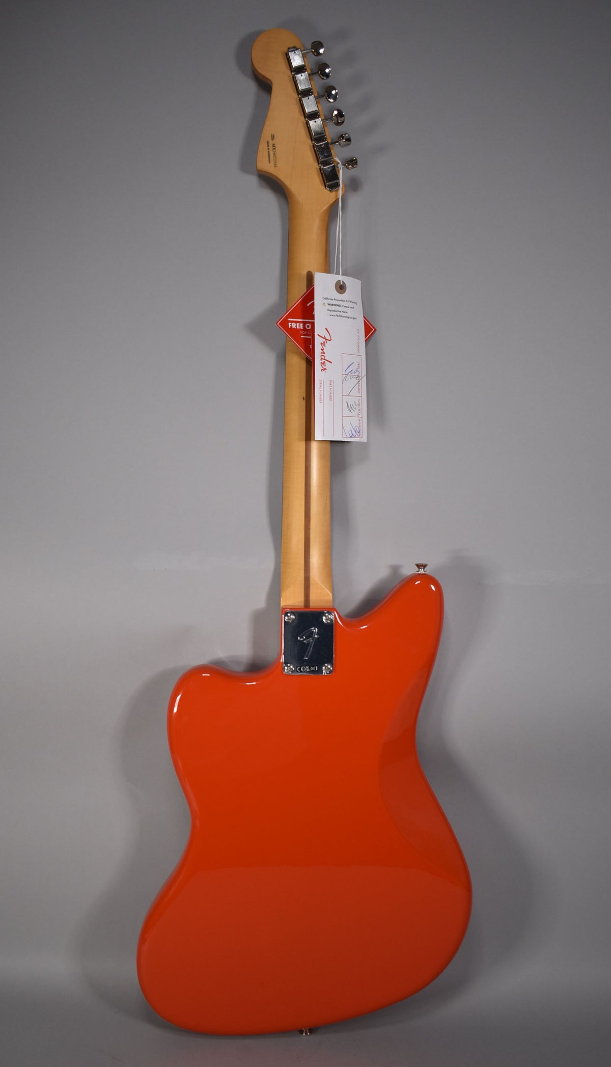 The back of a glossy coral red Jazzmaster with a light wood neck, chrome tuners, and a neck plate, with hang tags.