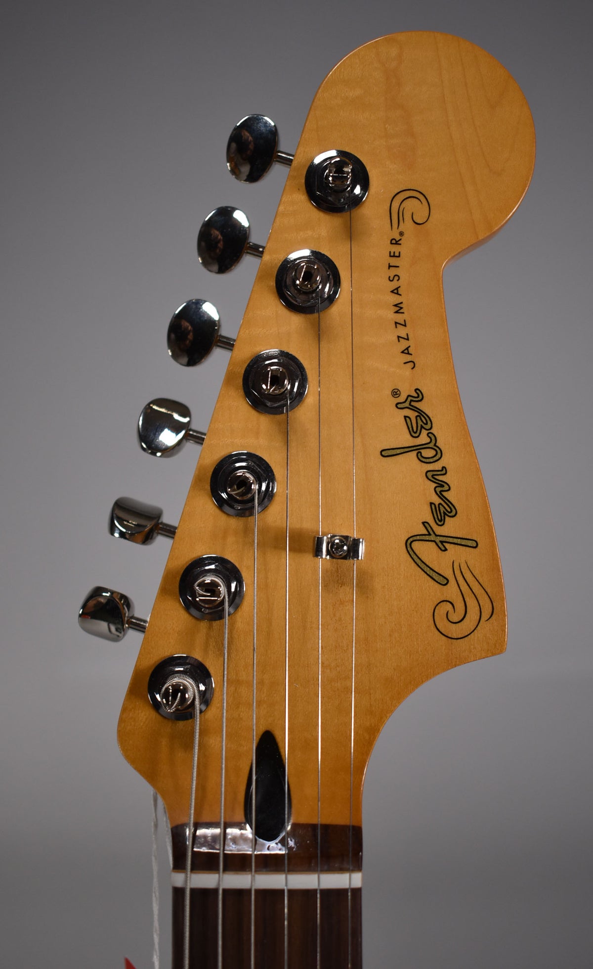 Close-up of the Jazzmaster's light wood headstock, angled left, showing six chrome tuners, a string tree, and the dark fretboard below.