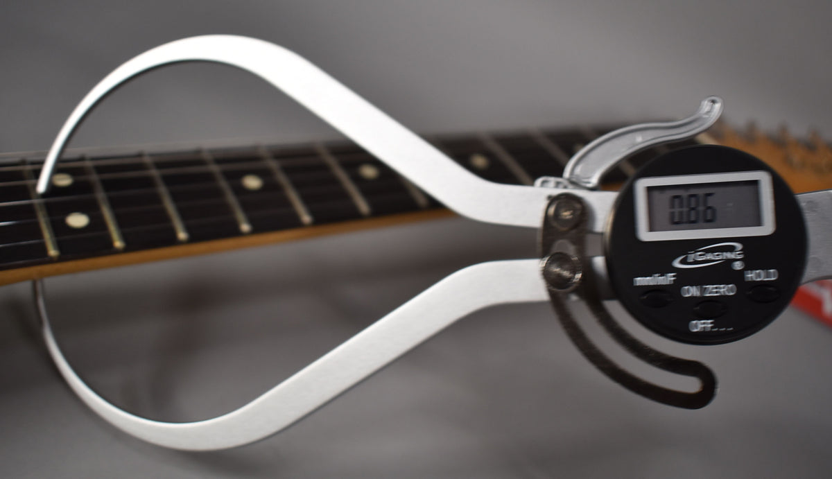 A digital caliper measures the fret height of the guitar's dark fretboard, showing "0.86" on its display.