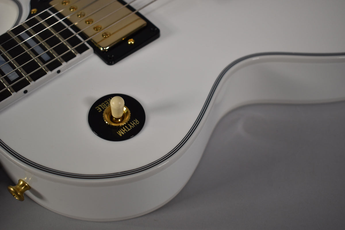 Close-up of a glossy white guitar body with black and white binding, showing a gold pickup, cream toggle switch, and gold strap button.
