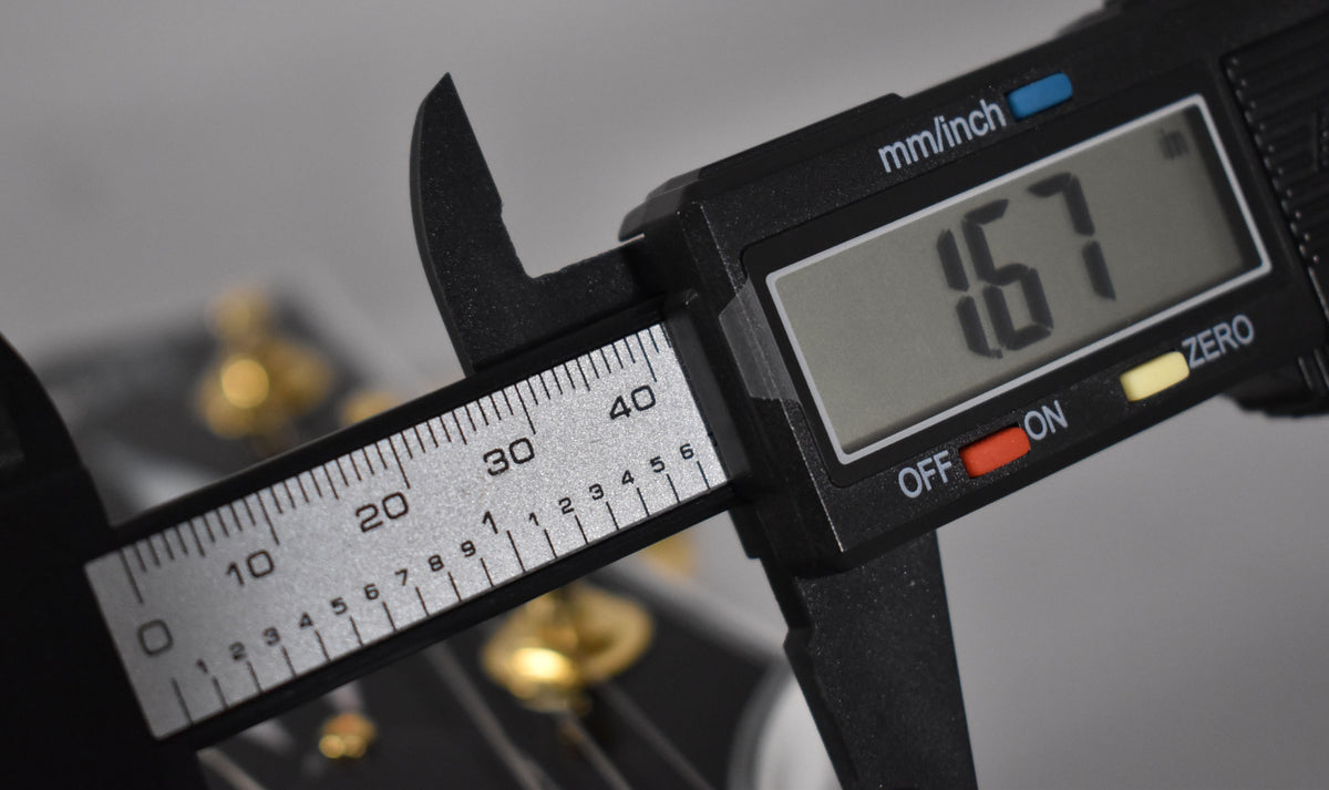 A digital caliper measures the nut width of the Les Paul, displaying "1.67" on its screen, with gold tuners blurred in the background.