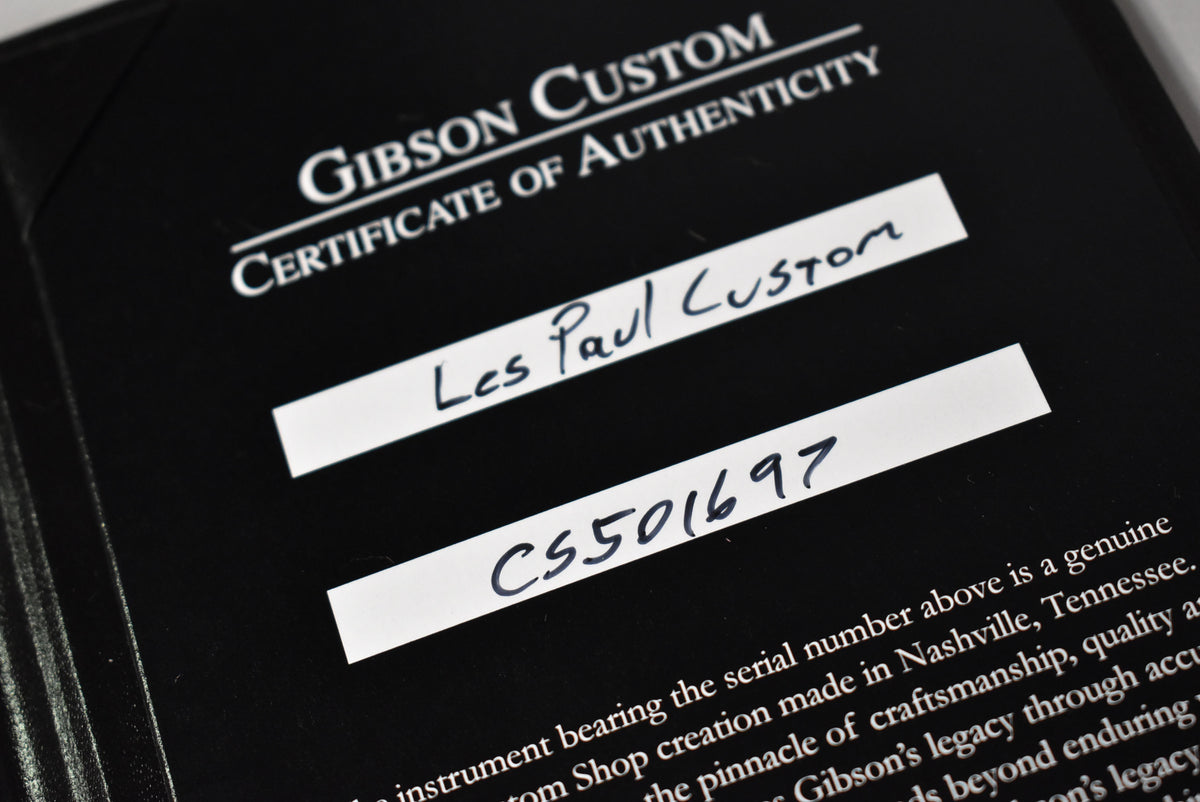 Close-up of a black Gibson Custom Certificate of Authenticity, showing "Les Paul Custom" and serial number "CS501697" handwritten on white labels.