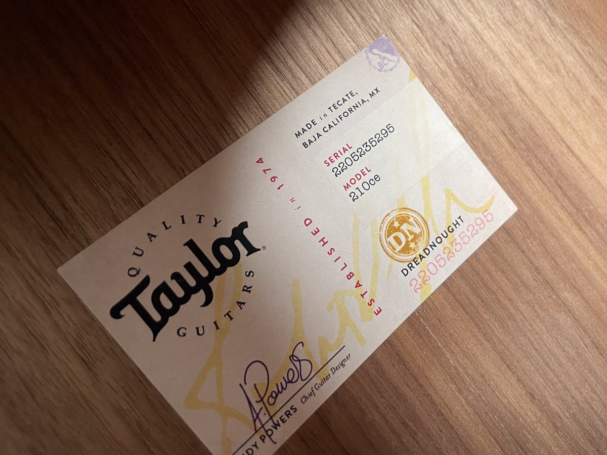 Close-up of a Taylor guitar spec sheet, angled on a light wood surface, showing serial number 2205235295 and model 210ce.