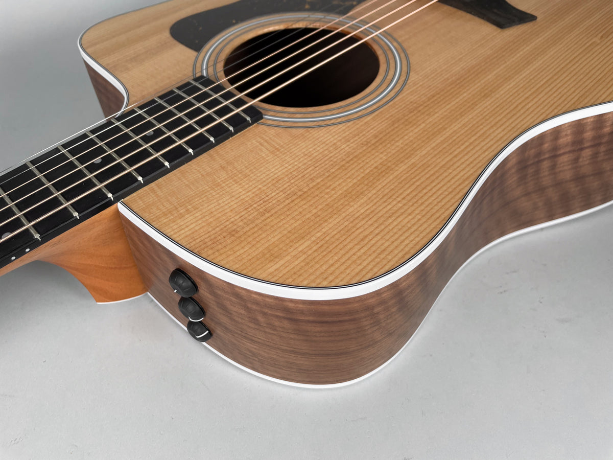 The upper body of a natural finish acoustic guitar, showing the soundhole, pickguard, and three black control knobs on the side.