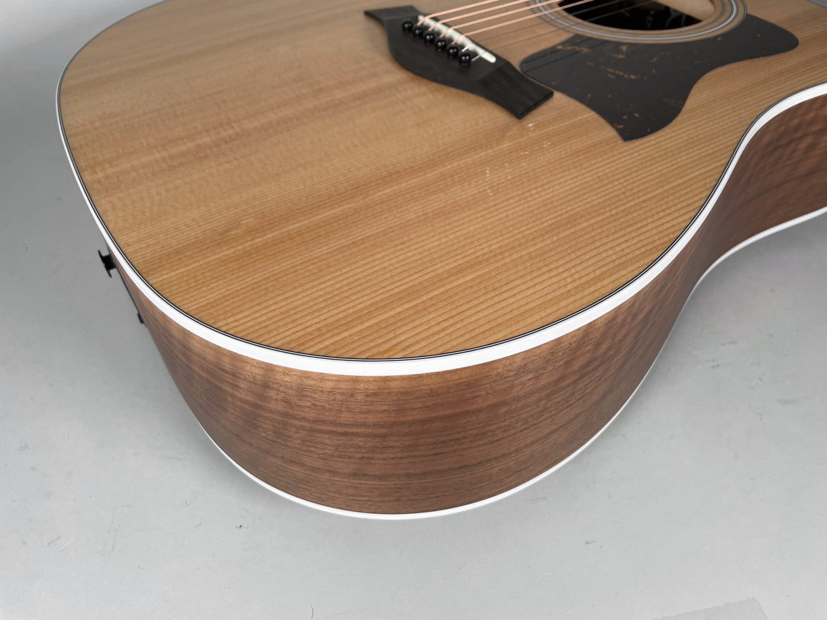 Upper bout of a natural finish acoustic guitar, showing light wood grain, dark pickguard, and white binding.