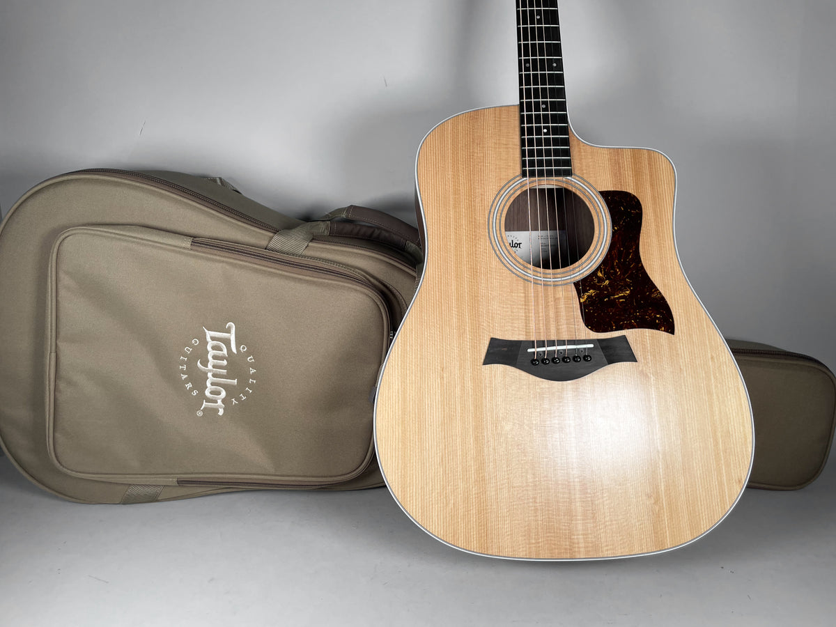 A natural finish acoustic guitar with a cutaway body and tortoiseshell pickguard, standing next to a tan soft case.