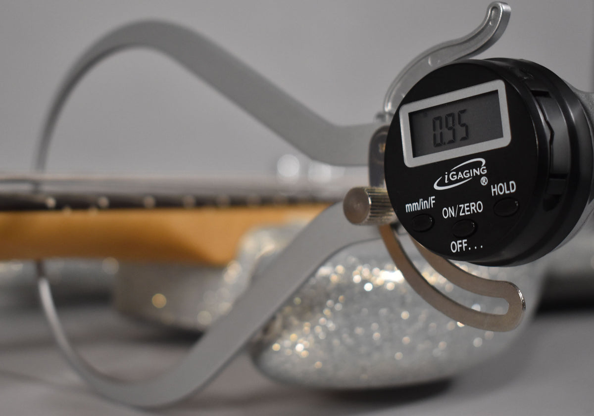 A digital caliper measures the bass's fretboard and strings, showing "0.95" on its display, with the sparkly body blurred below.