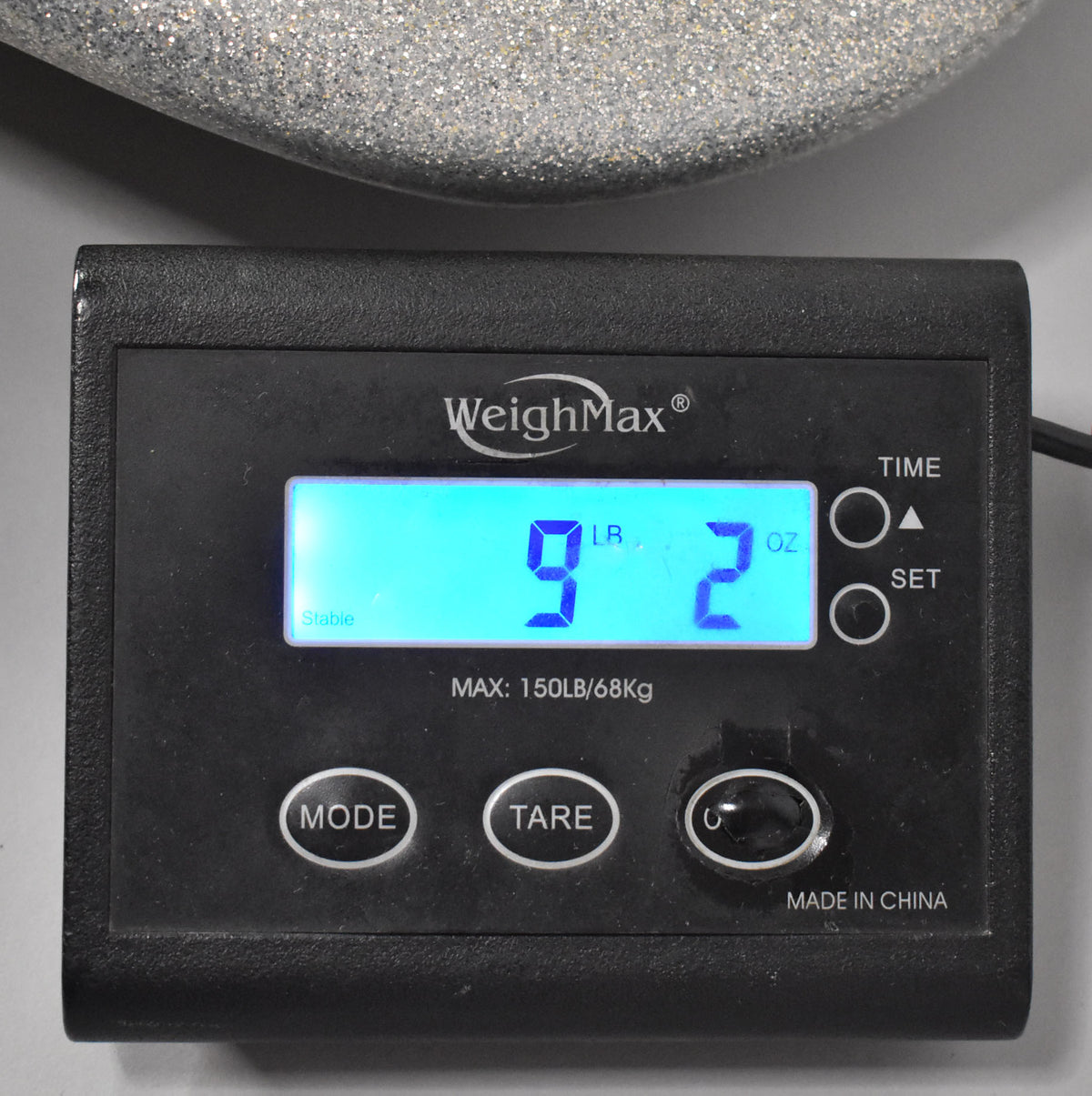 Close-up of a black digital scale displaying "9 LB 2 OZ" in blue, with a silver sparkle bass body resting on it.