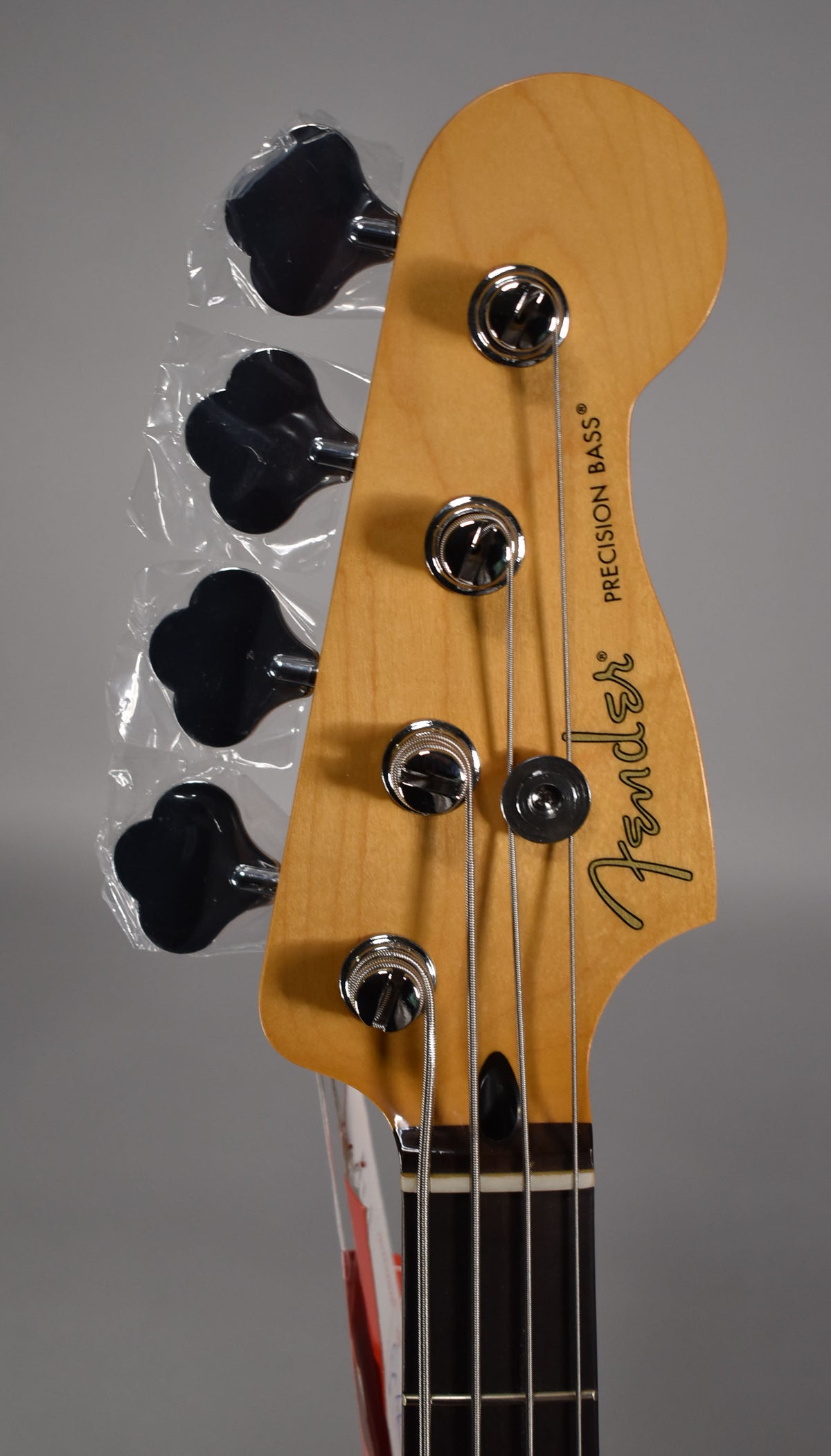 Close-up of the bass headstock, showing the light wood finish, chrome tuners, and black clover-leaf keys still in plastic.