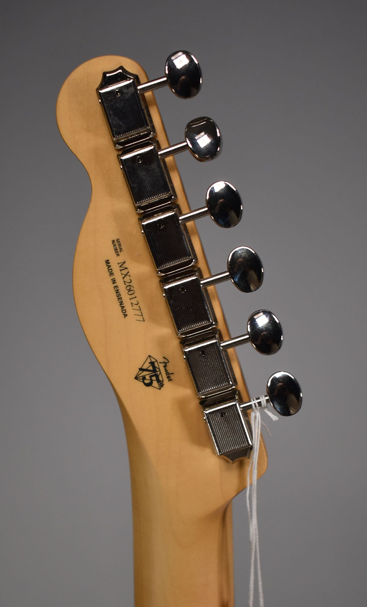 The back of a light maple headstock with six chrome tuners, angled slightly to the right, with a white tag tied to the lowest tuner.