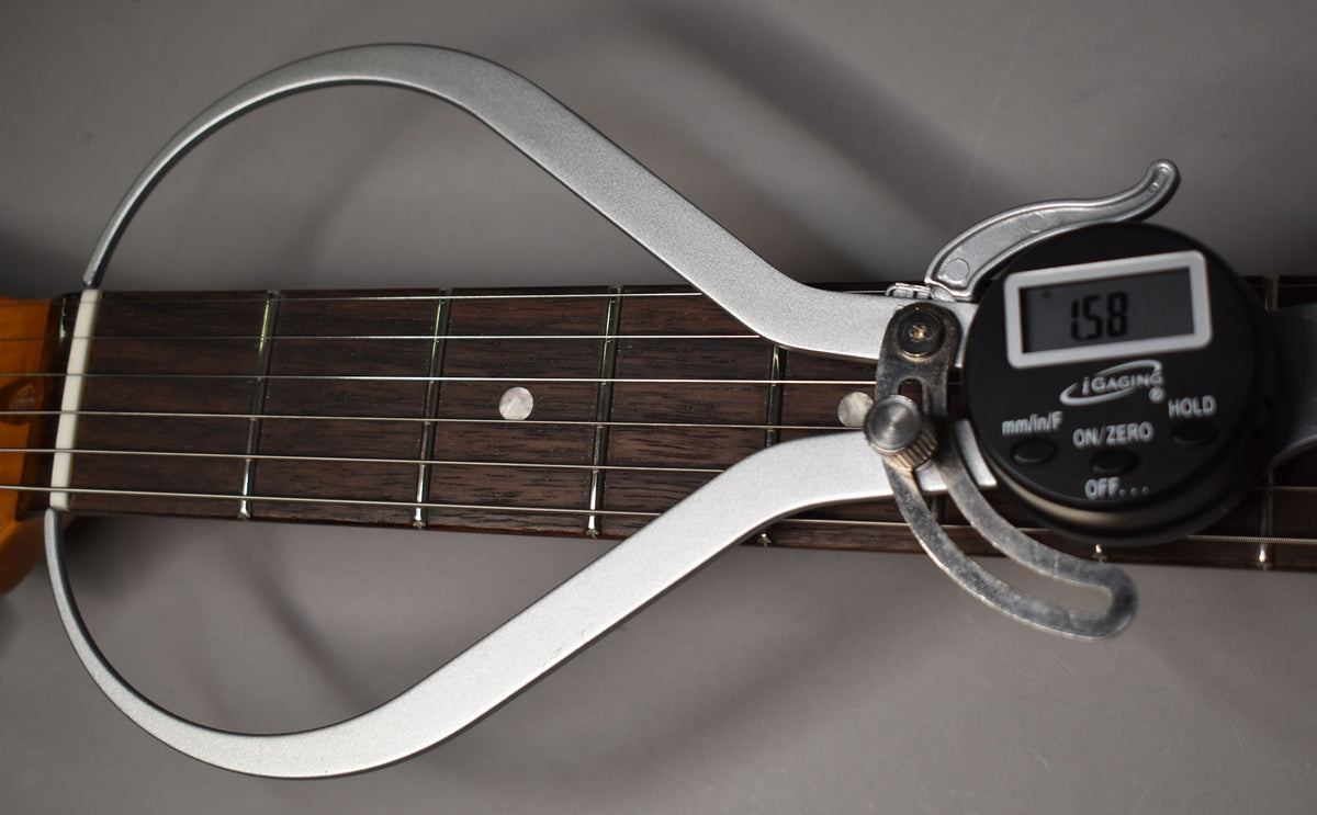 A digital fret height gauge measures the dark rosewood fretboard with white dot inlays and light strings.