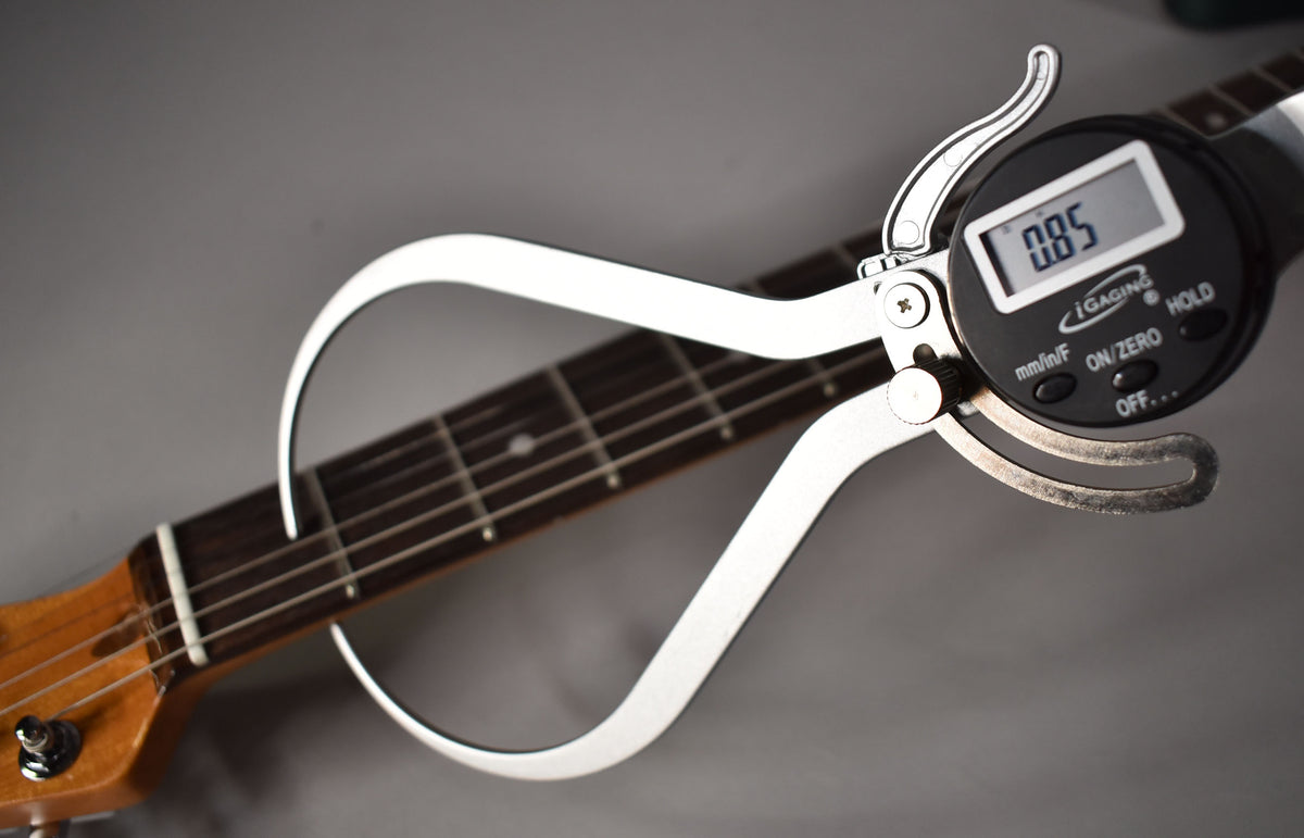 Close-up of the guitar's dark fretboard and light-colored headstock, with a digital caliper measuring the nut's thickness.