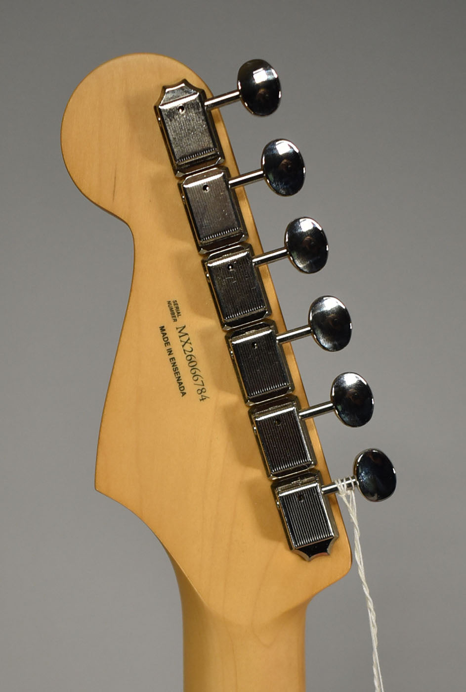 The back of a light maple headstock with six chrome tuners, showing a faint scratch near the top and a white string tag on the lowest tuner.