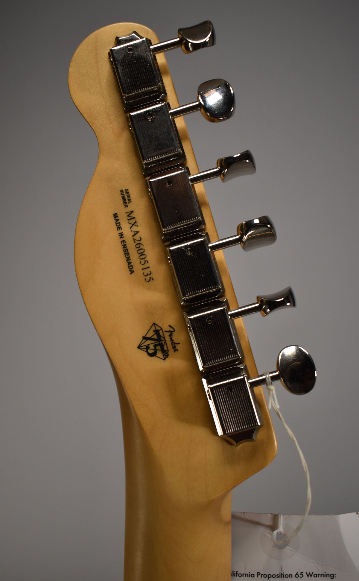 The back of a light wood guitar headstock with six chrome tuners and a white tag hanging from the lowest tuner.