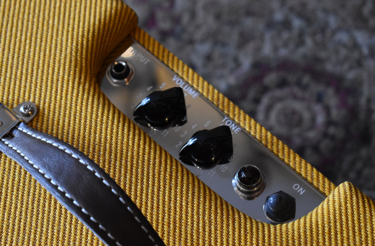 Close-up of the tweed amp's top, showing the shiny control panel with black Volume and Tone knobs, Input jack, and On switch.