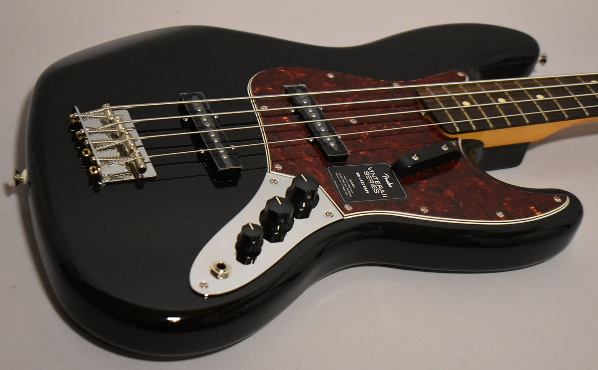The black gloss Jazz Bass body, with a tortoiseshell pickguard and chrome control plate, is angled from the lower right.