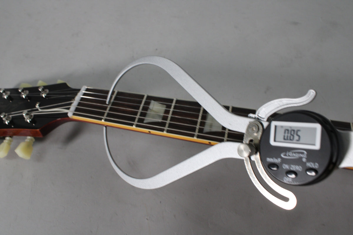 A digital caliper measures the guitar's fretboard at the nut, showing 0.85 on its screen, with the headstock and tuners visible.