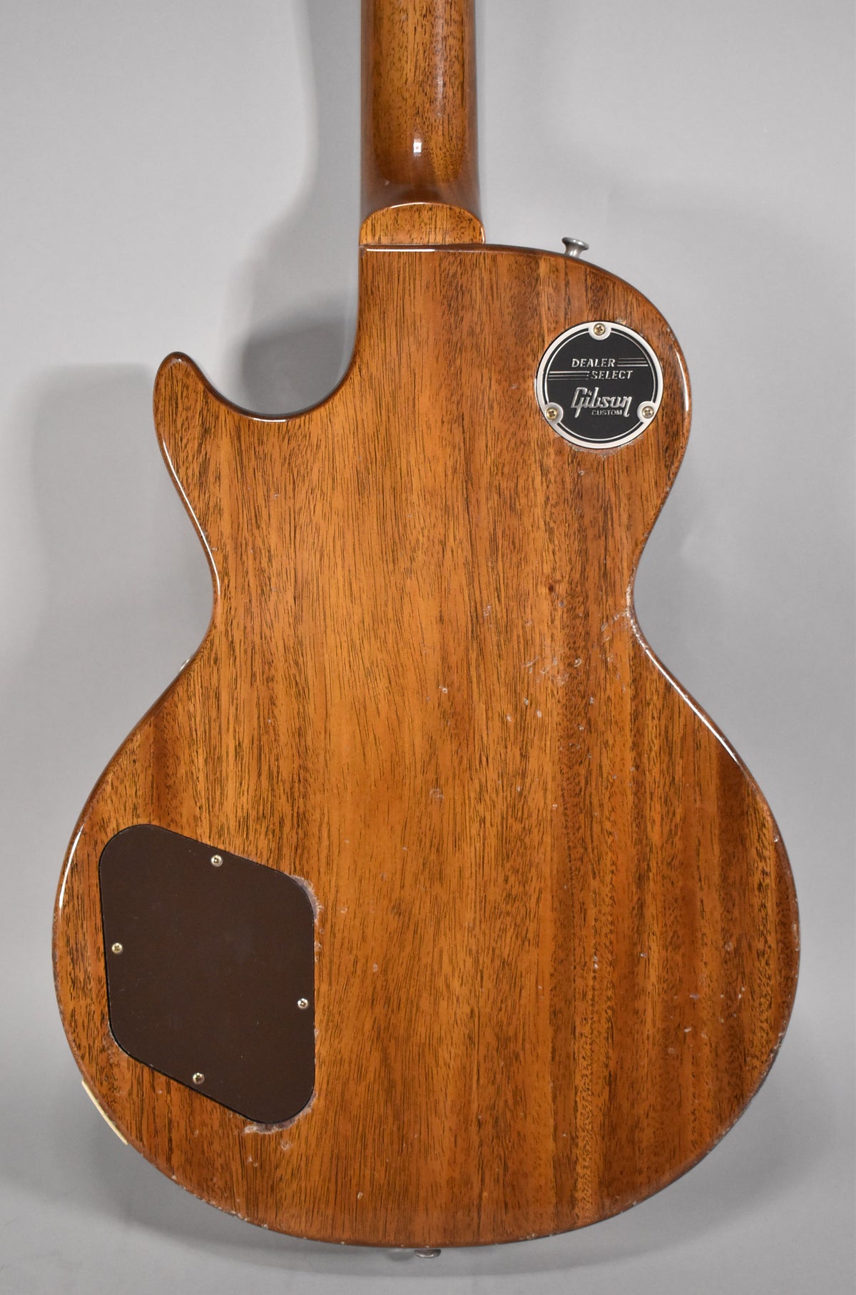 The back of a light-aged, brown wooden guitar with a black "Dealer Select Gibson Custom" plate and a dark brown control cavity cover.