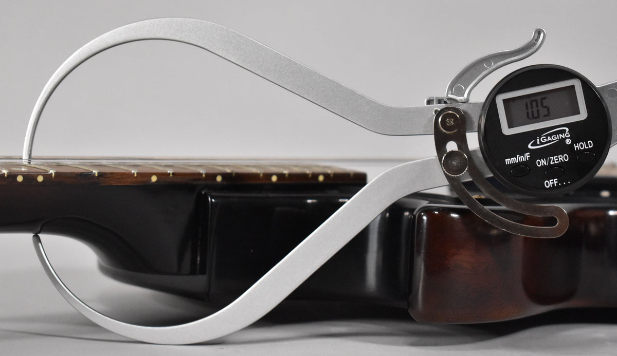 A digital caliper measures the neck thickness at the first fret, showing 1.05 inches, on the dark brown guitar.